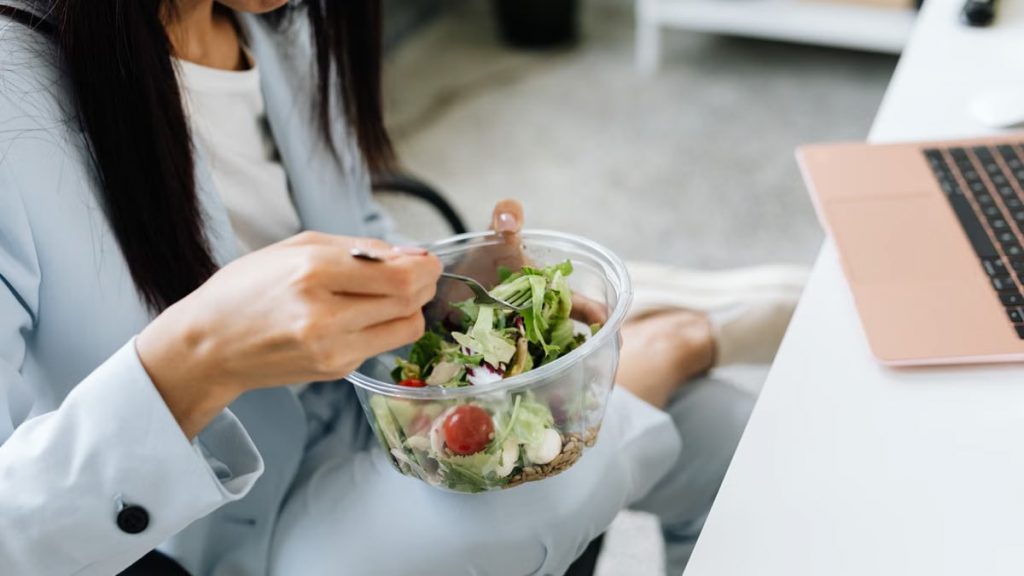 Adult woman eating a vegetable salad at her office desk, embodying healthy lunchtime habits.